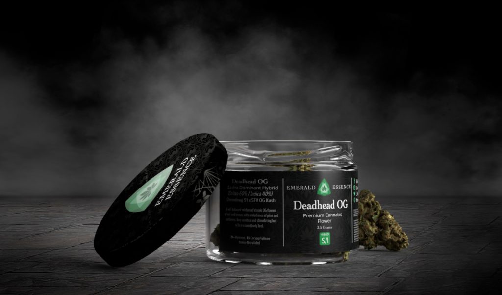 A glass jar with a black metallic lid and black matte label emblazoned with the emerald essence green emerald logo and strain information over a geometric pattern background. a cannabis flower lays nearby.